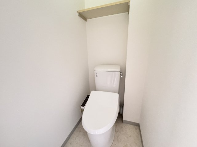 清潔感のある温水洗浄便座付きトイレです。白を基調とした明るい空間で、毎日気持ちよくご利用いただけます。上部には棚が設置されており、トイレットペーパーや掃除用品の収納にも便利。シンプルで使いやすい設計。