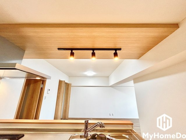 キッチンの天井部分にあしらわれた温もりある木目調クロスと、黒のライティングレールに配された暖色系スポットライトが、空間の洗練されたアクセントになっています。