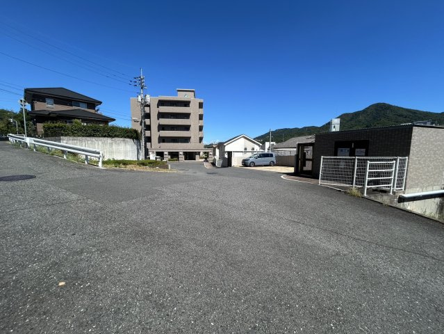 前面道路側から建物全体を確認できる現地写真です。周辺は低層の住宅を中心とした街並みで、背後には緑豊かな山並みも望めます。建物周辺は視界が開けており、マンションの位置や周辺環境をイメージしやすい立地です