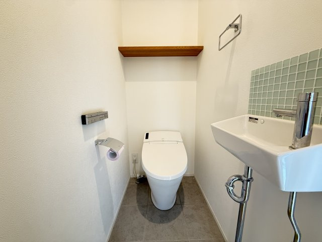 シンプルで清潔感のあるトイレ空間。手洗い器が設置されており使い勝手も良好です。上部には棚があり、トイレットペーパーや掃除用品の収納にも便利。コンパクトながら機能性をしっかり確保しています。　