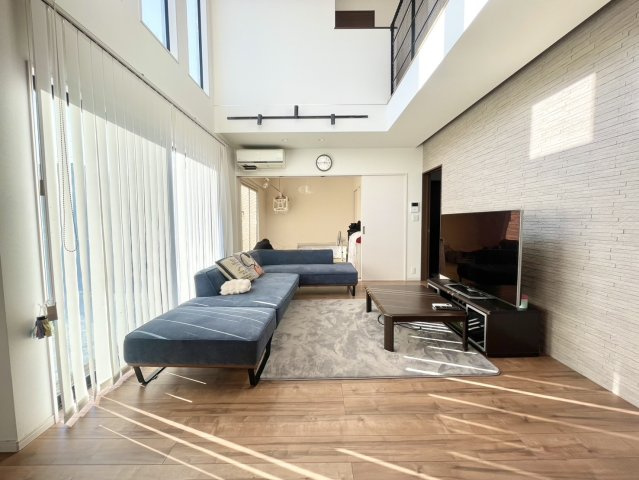 大きな吹き抜けとワイドな窓から自然光がたっぷり入り、1日中明るく開放感に満ちたリビング。木目の床と白い壁が調和し、上質で落ち着いた雰囲気。L字ソファを置いてもゆとりがあり、家族みんながくつろげる広さ。