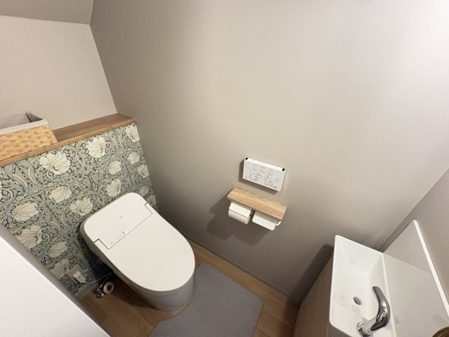 落ち着いた色合いの壁と、クラシカルな植物柄のアクセントクロスが調和したおしゃれなトイレ空間です。独立した手洗い器を備えており、使用後すぐに手を洗える便利な設計です。奥に棚も設けられています。