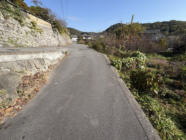 西側の公道はゆるやかにカーブした静かな生活道路で、車の通行も落ち着いた環境です。石垣のある風景が周辺の穏やかさを感じさせ、現地へのアクセスもスムーズです。