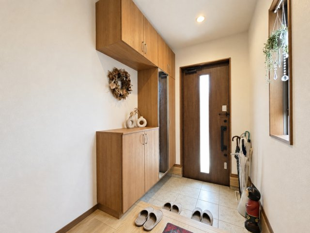 木目調の温かみある収納が並ぶ落ち着いた雰囲気の玄関です。大きめの下駄箱で収納力も十分。採光窓から光が入り玄関まわりが明るく清潔な印象に。季節の飾りつけも楽しめるスペースでお客様を心地よく迎えられます。