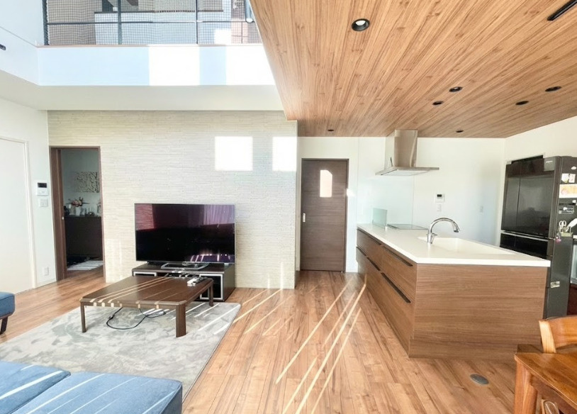 吹き抜けから明るい光が差し込む開放的なLDK。木目の天井と床が上質な温かみを生み、家族が自然と集まる居心地の良い空間。キッチンとダイニングの動線がスムーズで料理をしながら家族の様子が見守れる点も魅力。