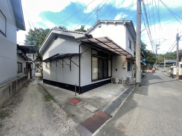 建物の外観と前面道路の様子。道路に面した立地で、周辺には戸建住宅が建ち並ぶ住宅街が形成されています。建物は1階部分が横に広がる造りとなっており、現地では建物の配置や周辺環境もあわせてご確認いただけます