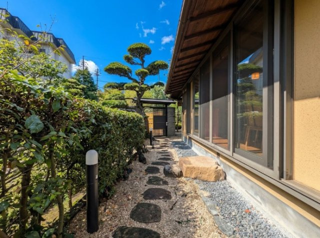 飛び石と砂利、丁寧に整えられた植栽が美しく調和した和のアプローチ。室内の大きな窓に沿って庭が広がり、住まいの中からも緑を身近に感じられます。青空と庭木の緑が映える、落ち着きと風情を感じさせる空間です。