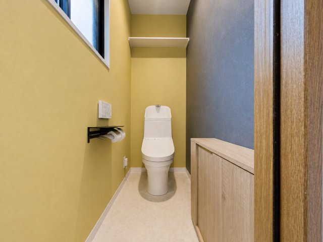 イエロー系と落ち着いた色合いのアクセントクロスを組み合わせた、おしゃれなトイレです。窓が設けられているため、自然光を取り込みやすく換気もしやすいのがポイント。手の届きやすい位置に棚も設けられています。