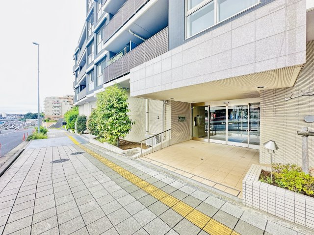 整備された広い歩道に面した明るいエントランス。　前面道路との間に十分なスペースがあり、ベビーカーの移動やお子様の飛び出し防止にも安心な設計です。フラットなアプローチで日々の出入りもスムーズに行えます。
