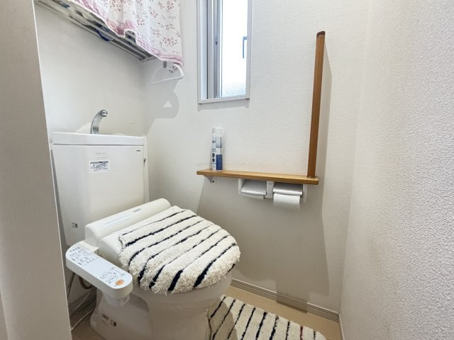 小窓付きで明るく清潔なトイレ空間。手すりと収納棚を備え、使い勝手にも配慮されています。温水洗浄便座付きで快適。花柄のカーテンがやさしい雰囲気を演出し、日常の中にも心地よさを感じられます。　