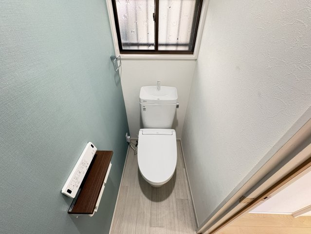 室内リフォーム済みで、清潔感のあるトイレ空間です。ブルーグリーン系のアクセントクロスが爽やかな印象を与え、シンプルながらもおしゃれな雰囲気に仕上がっています。背面には窓が設けられています。