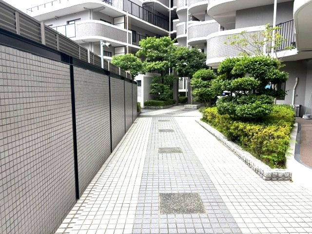 【美しく整備された敷地内アプローチ】上品なタイル敷きの通路が、マンション内へと優しく導きます。敷地内には青々とした植栽が丁寧に手入れされており、日々の移動の際にも緑豊かな心地よさを感じられます。