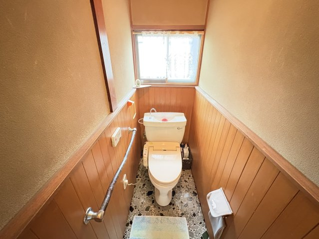 木の腰壁と明るい内装が調和し、落ち着いた雰囲気を演出するトイレ空間。手すり付きで高齢者の方も安心して使えるバリアフリー設計です。窓があることで自然光が入り、換気もしやすく、湿気対策にも安心。