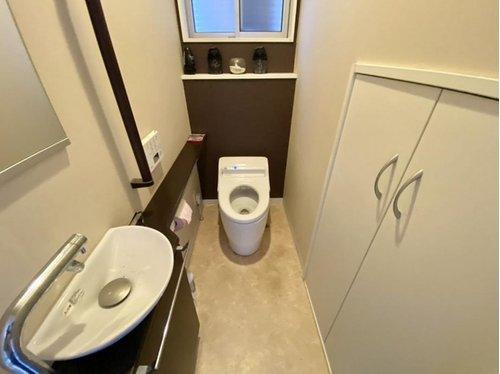 手洗い器付きの清潔感あるトイレ空間。明るいクロスと落ち着いたアクセントカラーで、快適な使用感を演出。収納棚も備わり、トイレットペーパーや掃除用品などもすっきり片付きます。