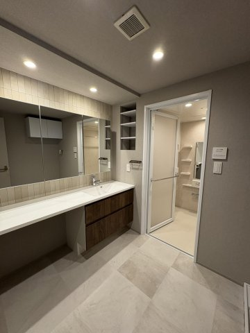 上質な素材感とデザイン性が美しく調和する洗面空間。洗練された間接照明の光が壁面のタイルを優しく照らし、床の大判タイルとともに気品ある雰囲気を演出します。奥の浴室へと繋がる、機能的で洗練された空間です。