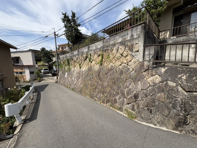 閑静な住宅街に位置し、周辺には戸建住宅が建ち並ぶ落ち着いた住環境です。道路面より高い位置に敷地があり、石積みの擁壁が特徴的な現地。周囲には緑も見られ、穏やかな雰囲気が感じられます。