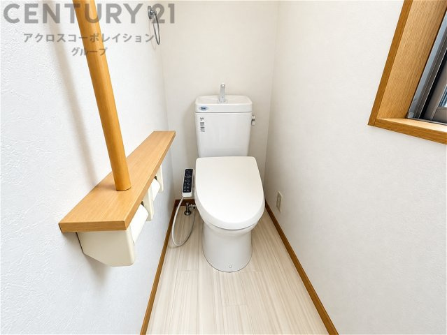 温水洗浄便座を備えた清潔感のあるトイレ空間。窓付きで自然光を取り込める明るい室内です。手すりや棚、ペーパーホルダーも設置され、毎日快適にお使いいただけます。