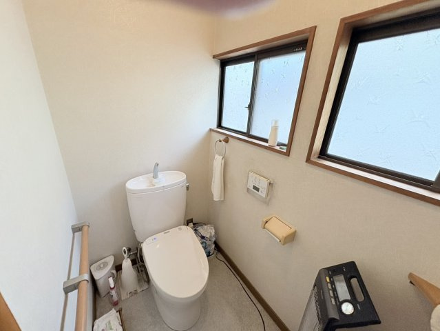 明るい窓付きのトイレ空間で、自然光が入り清潔感のある雰囲気です。温水洗浄便座付きで快適に利用できる設備。手すりが設置、動作のサポートにも配慮された造り。住居部分の設備として日常的に使いやすいトイレ。