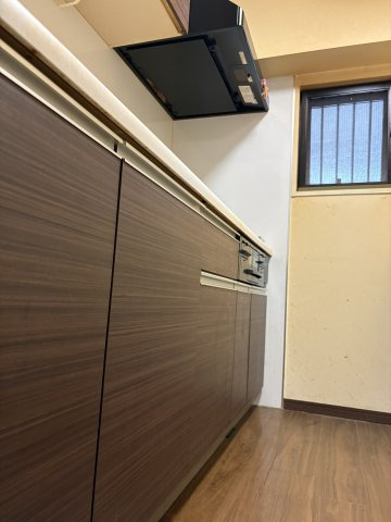 シックな木目調のデザインが美しいシステムキッチンです。奥には換気や採光に便利な窓が設置されており、調理中のにおいもこもりにくく風通し良好。足元まで落ち着いたフローリング調で統一されています。