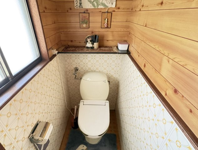 コンパクトにまとまった清潔感のあるトイレ空間。壁面のタイルや木目調の内装が落ち着いた雰囲気を演出しています。上部には小物を置ける棚もあり、実用性にも配慮。窓付きで換気もしやすく、快適。