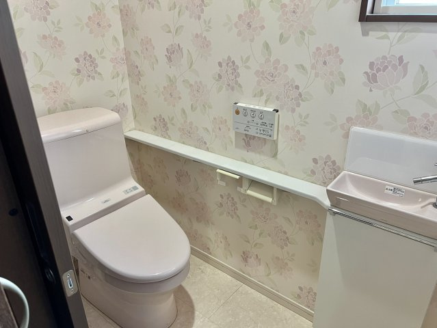 やさしい色合いのクロスが落ち着いた印象を与えるトイレ空間。手洗い器付きで来客時にも使いやすく、窓があるため自然光が入り換気もしやすい設計です。収納棚も備わり、見た目と実用性を両立しています。