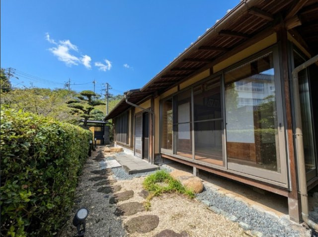 軒の深い日本家屋と庭が一体となった、趣のある景観が魅力。飛び石や砂利、植栽が建物に沿って配置され、和の風情を演出しています。大きな窓から庭を眺め、室内から四季折々の自然を身近に楽しめる住まいです。